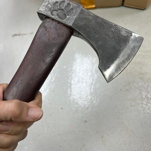 May include: A small, hand-forged axe with a dark wooden handle and a steel head. The head features a paw print design. The axe blade is sharp and ready for use. The axe is a tool for outdoor activities.