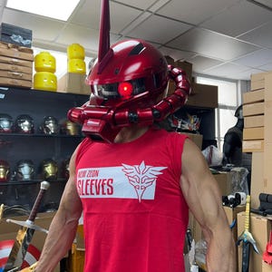 以下が含まれることがあります： 高いアンテナと赤く光る目を持つ、光沢のある赤いヘルメット。ヘルメットはコスチュームの一部です。その人は「NEW ZEON SLEEVES」という文字と白いエンブレムが入った赤いノースリーブのシャツを着ています。