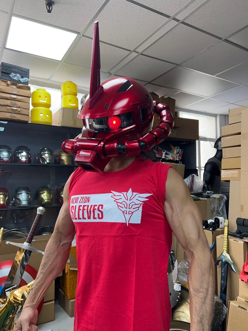 以下が含まれることがあります： 高いアンテナと赤く光る目を持つ、光沢のある赤いヘルメット。ヘルメットはコスチュームの一部です。その人は「NEW ZEON SLEEVES」という文字と白いエンブレムが入った赤いノースリーブのシャツを着ています。