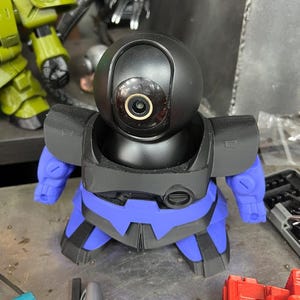 Puede incluir: Una c&aacute;mara de seguridad negra y azul con forma de robot, con una cabeza redondeada y una lente. La c&aacute;mara tiene brazos y piernas azules y un cuerpo negro. La c&aacute;mara est&aacute; sobre una superficie met&aacute;lica.