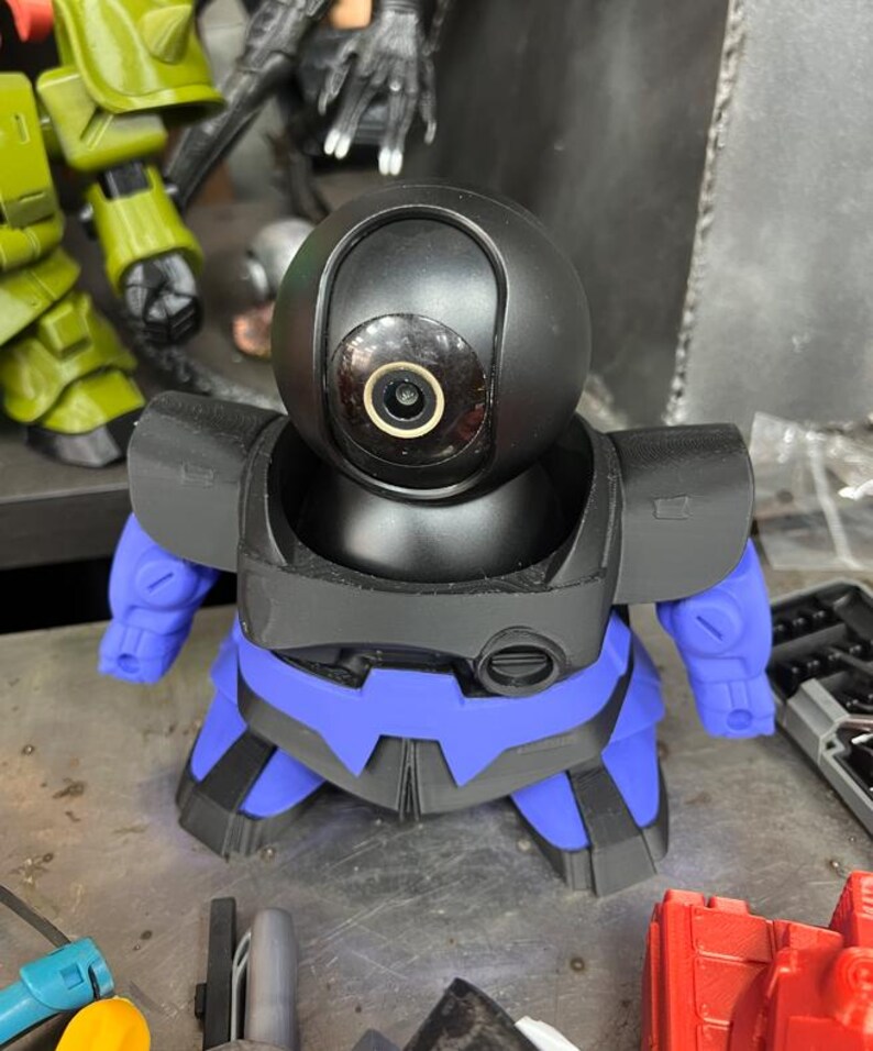 Puede incluir: Una c&aacute;mara de seguridad negra y azul con forma de robot, con una cabeza redondeada y una lente. La c&aacute;mara tiene brazos y piernas azules y un cuerpo negro. La c&aacute;mara est&aacute; sobre una superficie met&aacute;lica.