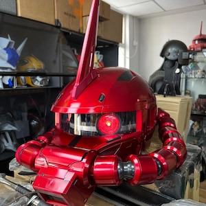 以下が含まれることがあります： 光沢のある赤いロボットヘルメット。目立つフィンと赤く光る目があります。ヘルメットは可動式のチューブと詳細な機械部品を備えており、未来的なテーマを連想させます。複雑で人目を引くデザインです。