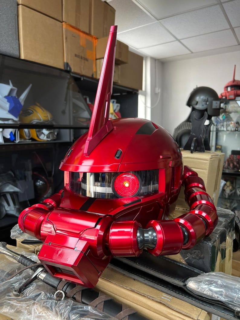 以下が含まれることがあります： 光沢のある赤いロボットヘルメット。目立つフィンと赤く光る目があります。ヘルメットは可動式のチューブと詳細な機械部品を備えており、未来的なテーマを連想させます。複雑で人目を引くデザインです。