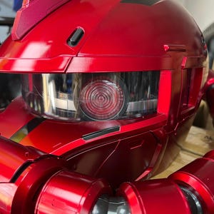 以下が含まれることがあります： 透明なバイザーと円形の詳細な目を持つ赤いロボットヘッドのクローズアップ。金属の表面は光を反射し、ロボットの頭部の複雑なデザインと鋭い角度を強調しています。ロボットの頭部は鮮やかな赤色です。