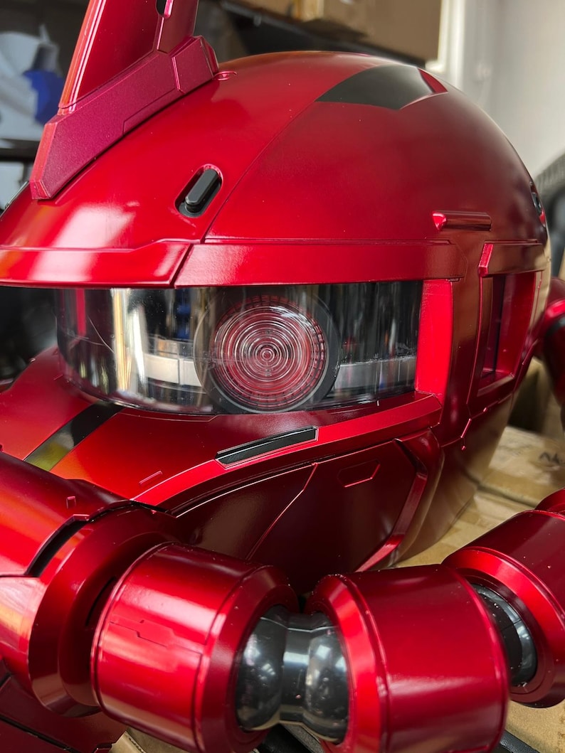 以下が含まれることがあります： 透明なバイザーと円形の詳細な目を持つ赤いロボットヘッドのクローズアップ。金属の表面は光を反射し、ロボットの頭部の複雑なデザインと鋭い角度を強調しています。ロボットの頭部は鮮やかな赤色です。