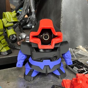 Puede incluir: Una peque&ntilde;a figura con forma de robot con una cabeza roja y negra, un torso negro y detalles azules. La figura tiene un ojo grande y circular y peque&ntilde;os brazos azules. El dise&ntilde;o sugiere un tema futurista o de ciencia ficci&oacute;n.