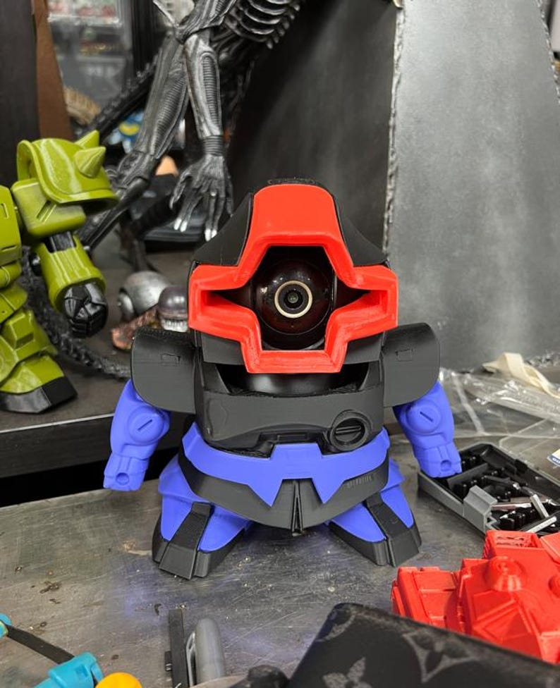 Puede incluir: Una peque&ntilde;a figura con forma de robot con una cabeza roja y negra, un torso negro y detalles azules. La figura tiene un ojo grande y circular y peque&ntilde;os brazos azules. El dise&ntilde;o sugiere un tema futurista o de ciencia ficci&oacute;n.
