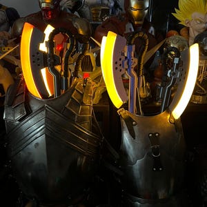 Op de afbeelding: Twee verlichte, metalen pantserstukken met gloeiende gele accenten. Het pantser heeft een donkere, verweerde uitstraling, met zichtbare klinknagels en riemen. De achtergrond bevat verschillende superheldenbeeldjes, waaronder Iron Man en Vegeta.