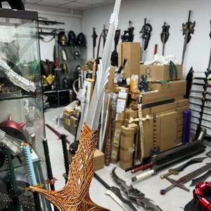 以下が含まれることがあります： 銀色の金属製ブレードと、柄の近くに白い葉のような装飾デザインが施された剣。柄は複雑な質感の銅色の格子模様で構成されており、幅の広い尖ったクロスガードのデザインへと広がっています。