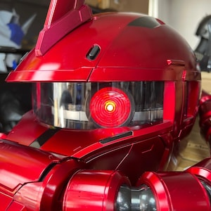 以下が含まれることがあります： 光沢のある赤いロボットヘルメットのクローズアップ。透明なバイザーと光る赤い目があります。ヘルメットは、洗練された未来的なデザインで、詳細な機械部品と上部のフィンを備えています。 全体的な美しさは、SFを連想させます。