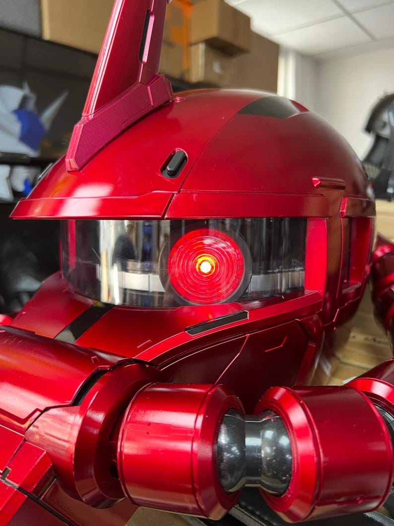 以下が含まれることがあります： 光沢のある赤いロボットヘルメットのクローズアップ。透明なバイザーと光る赤い目があります。ヘルメットは、洗練された未来的なデザインで、詳細な機械部品と上部のフィンを備えています。 全体的な美しさは、SFを連想させます。