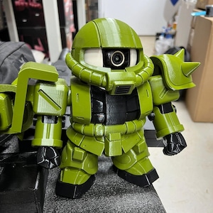 Puede incluir: Una figura de robot verde y negro con un ojo grande y único. La figura tiene un casco redondeado, hombros angulares y un cuerpo segmentado. El robot está en posición vertical, con los brazos a los lados.