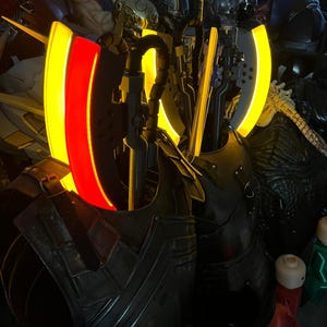 Op de afbeelding: Een verzameling gedetailleerde, donkergekleurde fantasy-pantserstukken en wapens. Het pantser heeft een metallic uitstraling met ingewikkelde ontwerpen. Sommige stukken hebben gloeiende gele en rode accenten, wat een opvallend visueel contrast cre&euml;ert. De items worden gepresenteerd in een zwak verlichte omgeving.