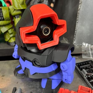 Puede incluir: Un juguete robot impreso en 3D con una combinaci&oacute;n de colores rojo, negro y azul. El robot tiene una cabeza distintiva con un marco rojo en forma de cruz alrededor de una lente negra. El cuerpo es negro y azul, con formas redondeadas.