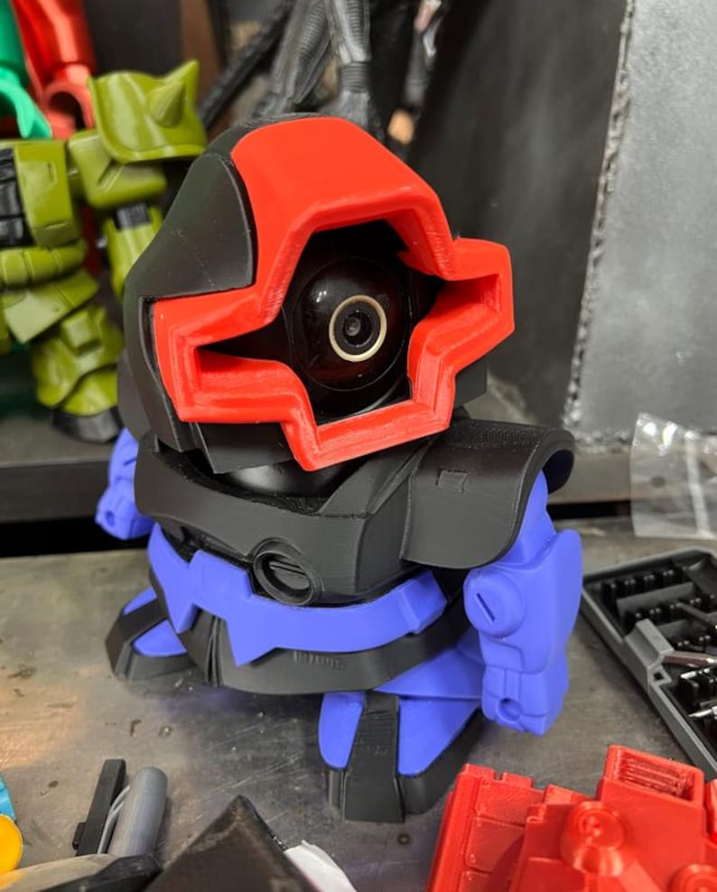 Puede incluir: Un juguete robot impreso en 3D con una combinaci&oacute;n de colores rojo, negro y azul. El robot tiene una cabeza distintiva con un marco rojo en forma de cruz alrededor de una lente negra. El cuerpo es negro y azul, con formas redondeadas.