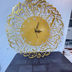 Könnte beinhalten: Eine goldfarbene Wanduhr mit arabischer Kalligraphie um das Zifferblatt. Die Uhr hat ein kreisförmiges Zifferblatt mit schwarzen Zeigern und Stundenmarkierungen. Die Uhr steht auf einer dunklen Oberfläche.