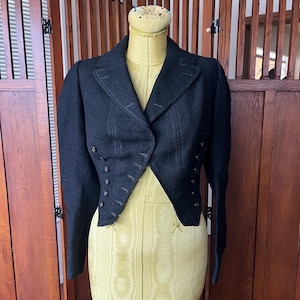 May include: A black wool jacket with a tailored fit, featuring a double-breasted design and decorative buttons. The jacket has a unique cut with a longer back and a shorter front, displayed on a mannequin.