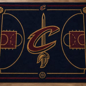 Puede incluir: Una alfombra rectangular con fondo azul oscuro y un diseño de cancha de baloncesto. El logotipo de los Cleveland Cavaliers está centrado en color vino y dorado. El texto Cleveland Cavaliers aparece a ambos lados, con iconos de balón en las esquinas.