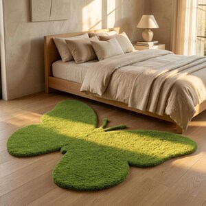 Puede incluir: Una alfombra verde brillante con forma de mariposa, hecha de un material suave y texturizado. La alfombra est&aacute; colocada sobre un suelo de madera clara frente a una cama con ropa de cama beige. La habitaci&oacute;n est&aacute; iluminada por luz natural.