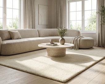 Peut inclure: Salon moderne avec un grand canapé d'angle beige, une table basse ronde en bois et un tapis crème. La pièce est baignée de lumière naturelle provenant de grandes fenêtres, et un olivier en pot ajoute une touche de verdure.