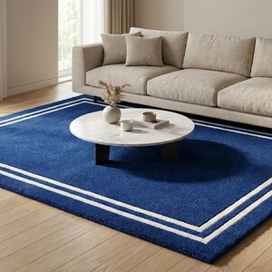 Puede incluir: Una alfombra rectangular azul oscuro con un borde blanco y un diseño de doble raya blanca. La alfombra está colocada sobre un suelo de madera clara, con un sofá beige y una mesa de centro redonda en el fondo. La habitación tiene una estética moderna y minimalista.