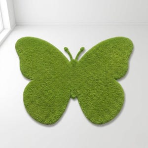 Puede incluir: Una alfombra verde con forma de mariposa con una superficie texturizada y afelpada. La alfombra es de un tono verde vibrante y est&aacute; colocada sobre una superficie blanca. El dise&ntilde;o presenta dos alas grandes y un cuerpo con antenas. Mide unos 150 cm de ancho.