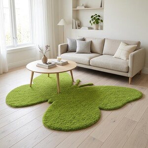 Puede incluir: Una alfombra verde brillante con forma de mariposa sobre un suelo de madera clara. La alfombra es el punto focal de una sala de estar, con una mesa de centro redonda de madera y un sof&aacute; beige en el fondo. Las alas de la alfombra son grandes y texturizadas.