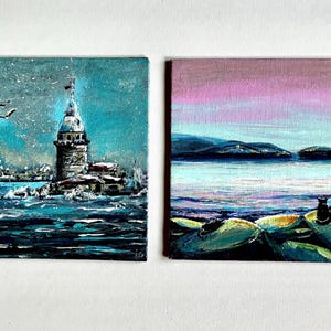 Könnte beinhalten: Zwei kleine quadratische Gemälde. Eines zeigt einen Turm in einem türkisfarbenen Meer mit Vögeln und Schnee. Das andere zeigt eine Seelandschaft mit einem rosa-violetten Himmel, Wasser und einer schwarzen Katze auf Felsen.