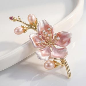 Puede incluir: Un broche dorado con forma de rama floreciente con pétalos de esmalte rosa, tres perlas ovaladas lisas y pequeños cristales transparentes en el centro de la flor. El diseño presenta un tallo delicado y una pequeña piedra rosa en la punta.
