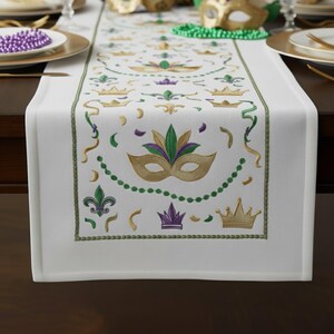 以下が含まれることがあります： 金、緑、紫のマルディグラのマスク、王冠、フルール・ド・リスのデザインが刺繍された白いテーブルランナー。 ランナーは緑色の縁取りで、濃い茶色のテーブルの上に置かれています。 装飾的なビーズとプレートが見えます。