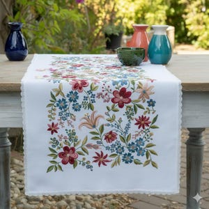 Op de afbeelding: Witte geborduurde tafelloper met een bloemenontwerp in rode, blauwe en perziktinten. De loper heeft een delicate kanten rand. Decoratieve vazen en een kom staan op de tafel.