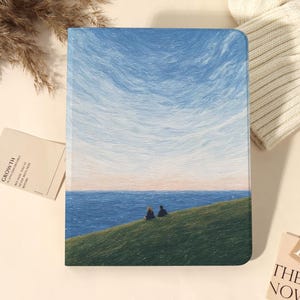 Könnte beinhalten: Eine rechteckige Tablethülle mit einem impressionistischen Gemälde eines grasbewachsenen Hügels, der über einen blauen Ozean unter einem texturierten Himmel blickt. Zwei kleine Figuren sitzen auf dem Hügel, umgeben von trockenem Gras und weißem Strickstoff.