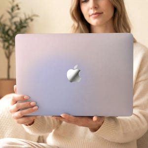 Puede incluir: Un portátil plateado con el logotipo de Apple se muestra. El portátil tiene un acabado degradado, que pasa del morado claro al plateado. El fondo está borroso, lo que sugiere un entorno suave y neutro.