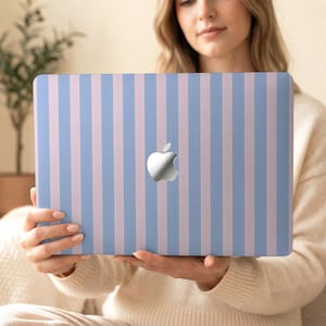 Könnte beinhalten: Ein Laptop mit einem hellblauen und rosa gestreiften Design. Der Laptop hat ein silbernes Apple-Logo in der Mitte. Das Design ist ein vertikales Streifenmuster. Der Laptop wird von einer Person gehalten.