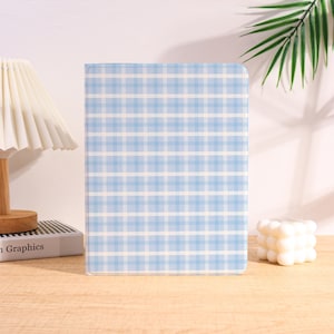 May include: A blue and white plaid notebook stands upright on a wooden surface. The notebook's cover features a light blue and white grid pattern. A small white candle and a lamp are visible in the background.