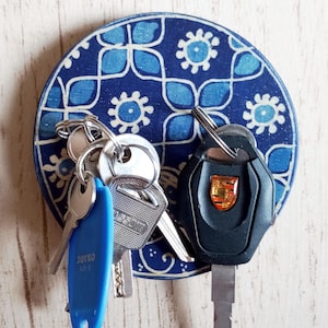 Può includere: Un portachiavi da parete circolare con un motivo floreale geometrico blu scuro e bianco. Il disco in legno sostiene diverse chiavi in metallo, tra cui una chiave per auto nera con uno stemma colorato e un'etichetta in plastica blu brillante.