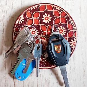 Könnte beinhalten: Ein runder Schlüsselhalter aus Holz mit einem rot-weißen Blumenmuster. Mehrere Schlüssel hängen daran, darunter ein blauer Schlüsselanhänger und ein schwarzer Autoschlüssel mit goldenem Emblem. Der Schlüsselhalter befindet sich auf einer hellen Holzoberfläche.