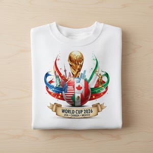 May include: White sweatshirt featuring a colorful graphic for the 2026 World Cup. The design includes a soccer ball with flags of the USA, Canada, and Mexico, along with the World Cup trophy and the text "WORLD CUP 2026 USA CANADA MEXICO."