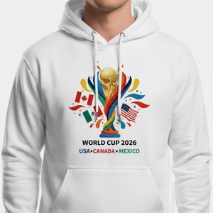 May include: White hoodie featuring a colorful graphic of the World Cup trophy, flags of the USA, Canada, and Mexico, and the text "WORLD CUP 2026 USA*CANADA*MEXICO". The design includes a gold trophy and vibrant accents.