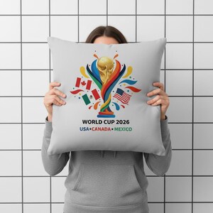 May include: A light gray square pillow featuring a gold trophy design with colorful accents and flags of the USA, Canada, and Mexico. The text "WORLD CUP 2026 USA CANADA MEXICO" is printed below the design.