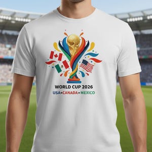 May include: White t-shirt featuring the World Cup 2026 logo with a golden trophy, colorful accents, and flags of the USA, Canada, and Mexico. The text "WORLD CUP 2026 USA CANADA MEXICO" is printed below the graphic.