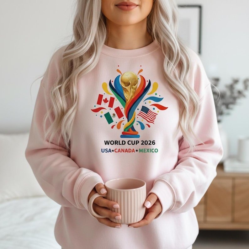 May include: A light pink sweatshirt featuring a colorful graphic for the 2026 World Cup, with the trophy and flags of the USA, Canada, and Mexico. The text "WORLD CUP 2026 USA CANADA MEXICO" is printed below the graphic.
