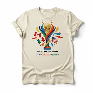 May include: Cream-colored t-shirt featuring a gold trophy design with colorful accents and flags of the USA, Canada, and Mexico. The text "WORLD CUP 2026" and the names of the host countries are printed below the graphic.