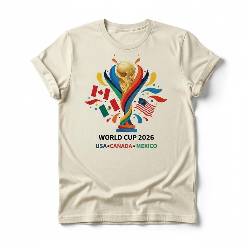 May include: Cream-colored t-shirt featuring a gold trophy design with colorful accents and flags of the USA, Canada, and Mexico. The text "WORLD CUP 2026" and the names of the host countries are printed below the graphic.