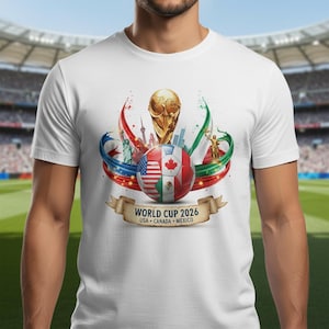 May include: White t-shirt featuring a colorful graphic design for the 2026 World Cup. The design includes a soccer ball with flags of the USA, Canada, and Mexico, along with the World Cup trophy and the text "WORLD CUP 2026 USA CANADA MEXICO".