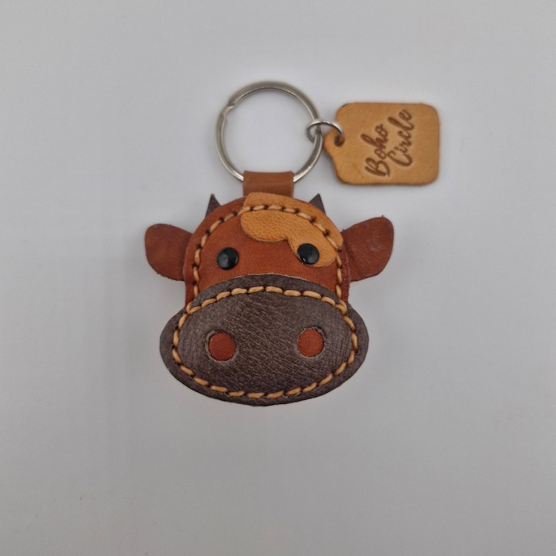 May include: A handcrafted leather cow keychain with a metal ring. The keychain features a brown and tan cow face with stitched details and black eyes. A small leather tag with the text "Boho Circle" is attached.