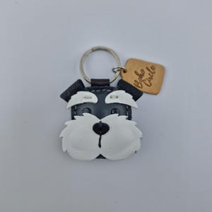 Pu&ograve; includere: Un portachiavi in pelle nera, bianca e marrone chiaro a forma di muso di cane Schnauzer. Il portachiavi ha un anello in metallo argentato e un piccolo cartellino in pelle marrone chiaro con la scritta "Boho Circle".