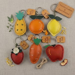 May include: A collection of six leather fruit and vegetable keychains including a pineapple, orange, lemon, strawberry, carrot, and apple. Each item features hand-stitched details, a metal ring, and a small tag labelled Boho Circle.