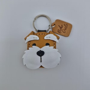Pu&ograve; includere: Un portachiavi a forma di cane in pelle fatto a mano, con corpo marrone, dettagli bianchi e naso nero. Il portachiavi ha un anello argentato e un piccolo tag in pelle con la scritta "Boho Circle".
