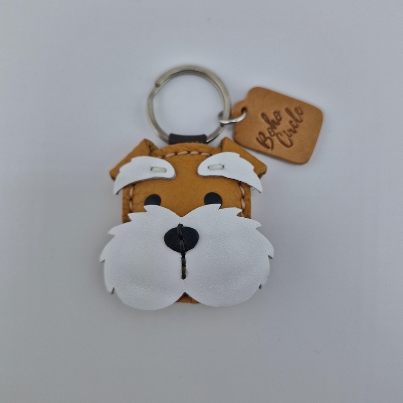 Pu&ograve; includere: Un portachiavi a forma di cane in pelle fatto a mano, con corpo marrone, dettagli bianchi e naso nero. Il portachiavi ha un anello argentato e un piccolo tag in pelle con la scritta "Boho Circle".